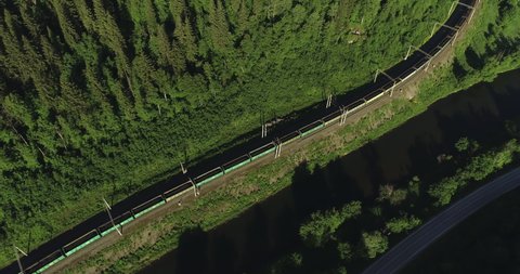 Freight train with coal carriages an electric locomotive by two-sided Siberian railways near river in the forest mountains / Aerial drone mid view at summer sunset