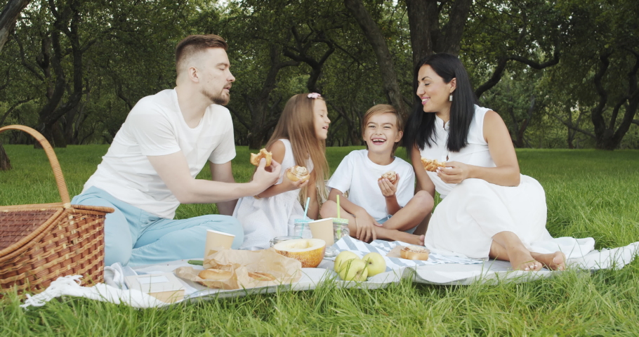 Happy family with children relaxing on the grass and eating delicious buns during a picnic in the green garden.