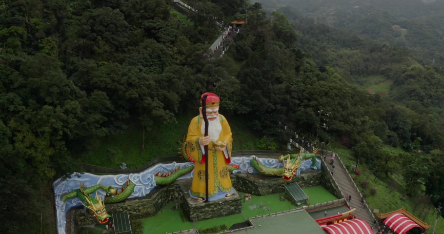 Decorative elderly religious male statue on temple rooftop. Aerial view rising along lush mountain pathway to ornate temple building, Taipei, China.
