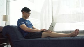 young asian fit man in sportswear sitting on sofa watching video training fitness online tutorials on laptop and exercising with dumbbells in living room at home . sport male workout in quarantine 