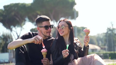 Portrait young couple of tourists sitting in sunglasses in the park on the lawn. Eat ice cream cones. Cheerful friends feasting on delicious ice cream on an urban background. Rome, Italy