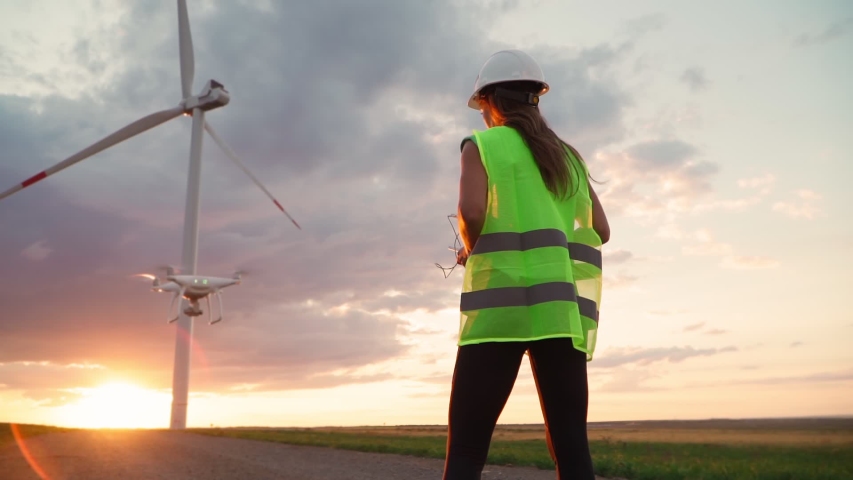 Woman Ecology specialist in uniform and helmet holding joystick controlling flying drone working at windmill on beautiful sunset background. Modern technology. Slow motion. Royalty-Free Stock Footage #1057037909
