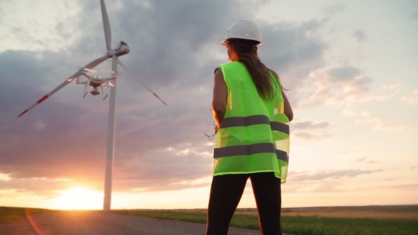 Woman Ecology specialist in uniform and helmet holding joystick controlling flying drone working at windmill on beautiful sunset background. Modern technology. Slow motion. | Shutterstock HD Video #1057037909