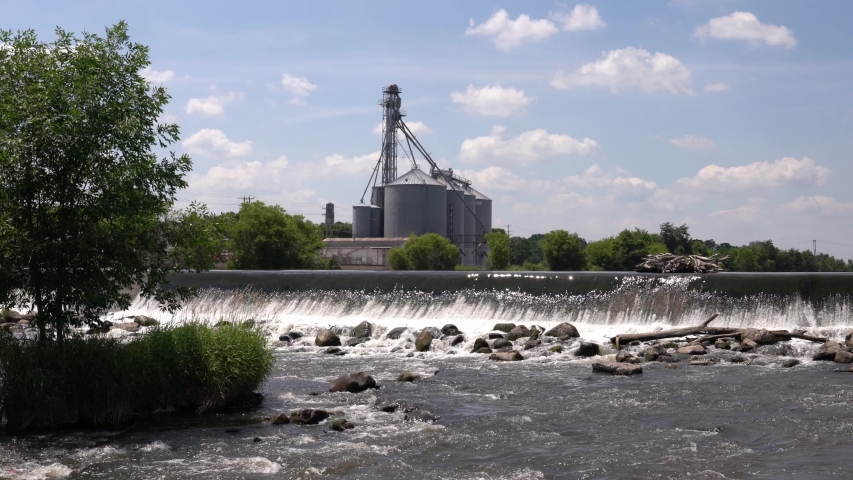 Water cascades or waterfalls down a concrete dam near Echo park and Echo lake where the White River flows into the Fox River in Burlington Wisconsin with a grain silo or elevator in background.