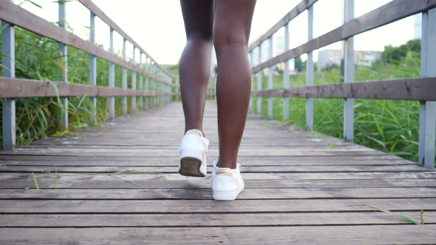 young African-American woman with long slim legs in white comfortable sneakers walks along wooden bridge among green reeds backside view Royalty-Free Stock Footage #1057043960
