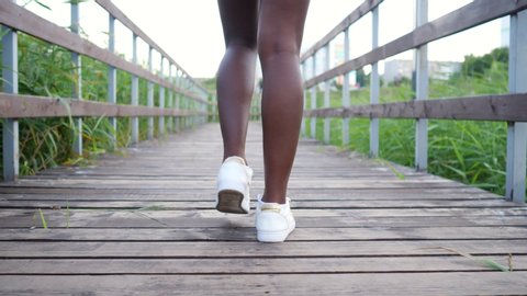 young African-American woman with long slim legs in white comfortable sneakers walks along wooden bridge among green reeds backside view
