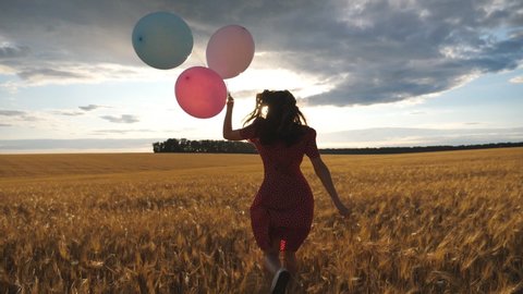 Rear view of happy girl in red dress running through golden wheat field with balloons in hand at sunset. Young woman with brown hair having fun while jogging among barley meadow. Freedom concept