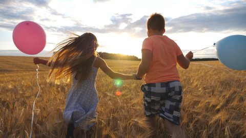 Couple of little kids with balloons running through wheat field, turning to camera and smiling. Small girl and boy holding hands of each other and jogging among barley meadow. Concept of child love