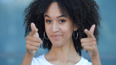 Attractive African American teen girl with makeup and afro hairstyle points her fingers at the camera, smiles toothy. Young ethnic woman looks approvingly, you are right, good and successful idea!