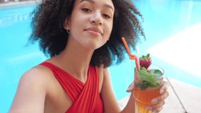 Close up of happy face young Afro blogger girl speaking with spectators viewer or friend online, sitting in luxury poolside, drinking ice fruit cocktail with mint and making photo or video network