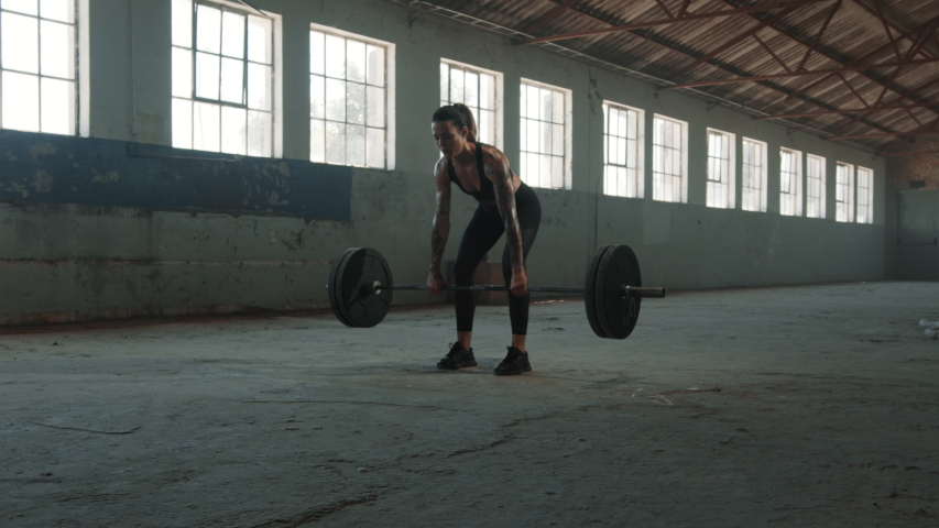 Fit woman performing the deadlift workout with barbell in old warehouse. Strong female athlete with muscular body lifting weights.
