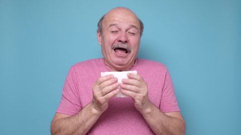 Infected man blowing his nose in tissue paper because of being ill isolated on blue background
