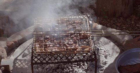 Backyard charcoal barbecue/braai with meat (pork sausages and lamb chops) secured on a metal grid. The prepared meat is placed in a metal dish using braai tongs