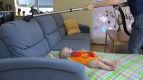 Cute newborn baby in orange body cloth lies on sofa as housewife woman cleans dust with vacuum cleaner. 4K UHD video clip.
