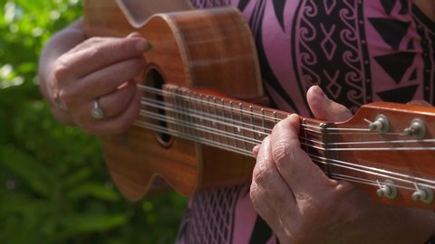 Close up of local woman playing double string Ukulele while wearing a pink Polynesian print dress. 