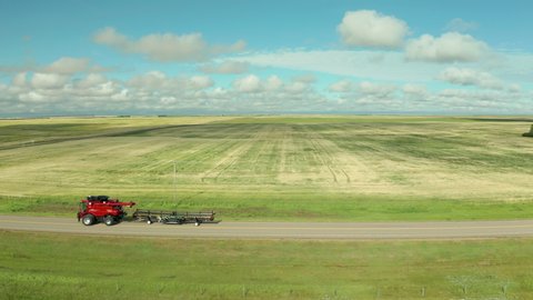 Modern red combine harvester versatile machines drive on dirt roads by expansive flat green farmland of crops on sunny day, Saskatchewan, Canada, above aerial sideways