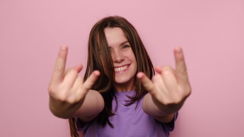 Portrait of cheerful young woman makes rock n roll sign, yells loudly, look at camera, wears purple t-shirt, devil horns hand sign gesture, stands over pink studio background, feels self confident