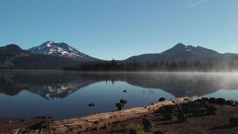 Aerial drone flight over log amazing reflection water lake two mountains broken top south sisters cascade forest range blue sky fog mist beautiful sparks lake central bend oregon pacific northwest usa