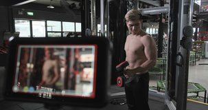 Sportman athlete doing exercise with dumbbells to camera while recording tutorial video of fitness training in modern gym. His muscular athlete showing shooting webinar video and making vlog.