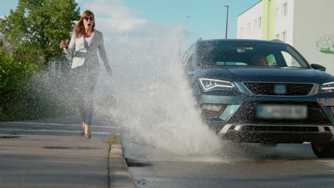 SLOW MOTION, LOW ANGLE: Unsuspecting businesswoman walking along sidewalk gets splashed by careless driver driving down the empty road. Senior woman drives into a puddle, splashing water at woman