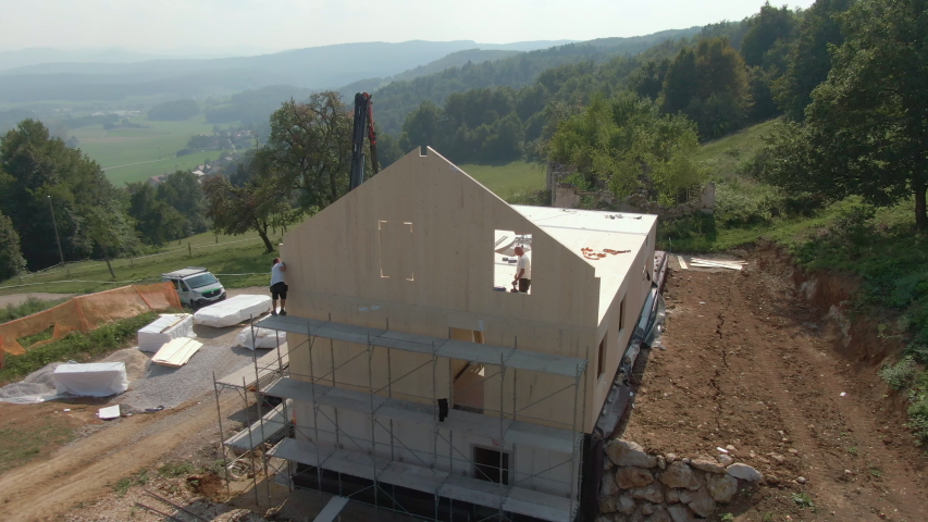 DRONE: Flying around a construction site of a modern sustainable house as truck boom lifts a CLT panel up to a floor full of contractors at work. Aerial view of a sustainable house under construction Royalty-Free Stock Footage #1057085990