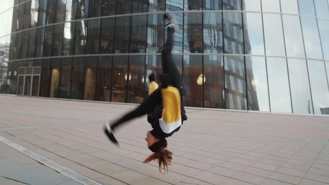 Young sporty woman performing aerial cartwheel while practicing parkour outdoors in front of modern glass building in the city