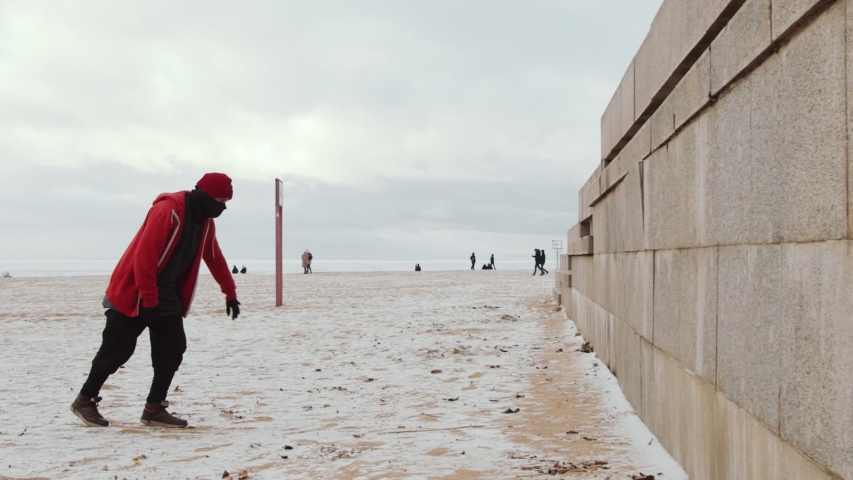 Slow motion shot of young man in sportswear and neck warmer running straight up the wall and doing backflip while practicing parkour outdoors on winter day Royalty-Free Stock Footage #1057089050