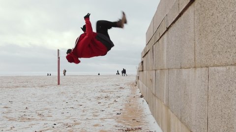 Slow motion shot of young man in sportswear and neck warmer running straight up the wall and doing backflip while practicing parkour outdoors on winter day