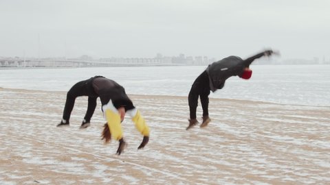 Slow motion shot of young athletic man and woman in sportswear performing cartwheel and backflips together while doing parkour on snowy lakeshore on winter day