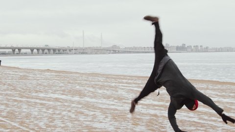 Slow motion shot of professional male athlete performing cartwheel and four backflips in a row while doing parkour outdoors on snowy shore of frozen lake