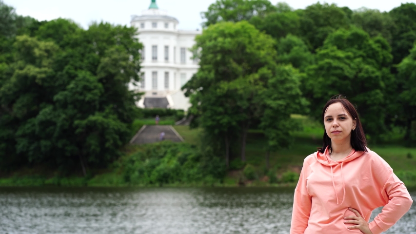Portrait of young woman posing near lake in summertime. Female tourist standing and looking at camera on blurred background of pond and estate.