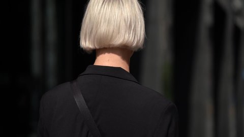 A transgender young girl with short white hair walks on a street in the city, sitting from the back, then turns around and poses, wearing a black jacket and a shoulder bag