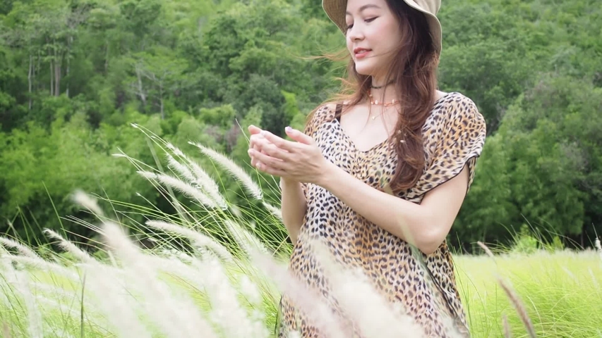 Asian woman in leopard pattern dress standing in blowing grass flower field with mountain in background.
