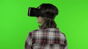 Young woman using VR app headset helmet to play simulation game. Watching virtual reality 3d 360 video. Isolated on chroma key green background in studio. Girl in VR goggles looking around