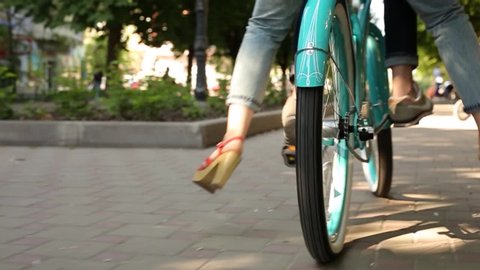 Back view of man pedalling a bike riding with woman on a seat or cargo rack. Low angle of young couple feet riding on bicycle together in park. Romantic boyfriend cycling with girlfriend. Slow motion.