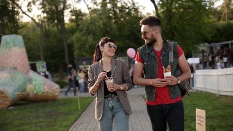 Stylish hipster couple walking eating ice cream and waffles laughing in city park. Romantic young man and woman having fun outdoors on their leisure. Boyfriend and girlfriend in slow motion.