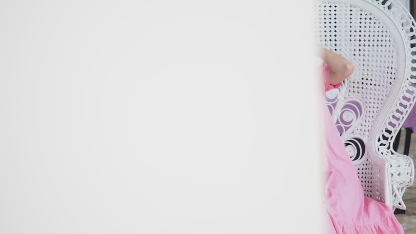 Asian woman in pink dress sitting on classic chair on balcony near tea table in afternoon. Female looking away.