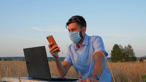 business men at remote work. A young man wearing a Covid-19 mask works for a laptop in a wheat field.
