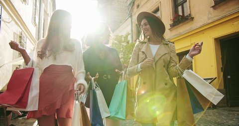 Beautiful mixed-races happy girlfriends in good mood walking on street with shopping bags and talking. Beautiful joyful females walking in city with purchases after shopping. Shopaholic concept