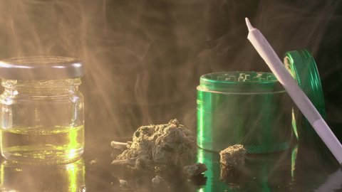 Close up of a medical marijuana joint a heap of weed crusher and cannabis oil on black background surrounded by haze shot in 4k super slow motion
