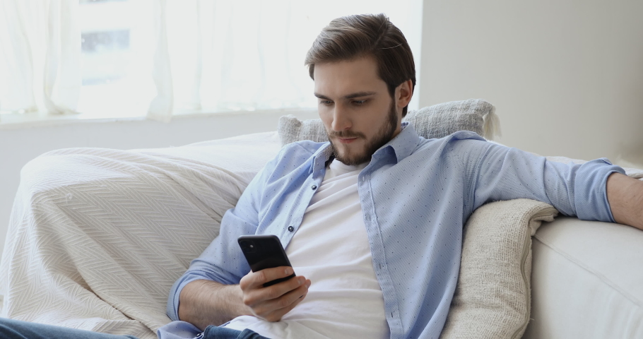 Handsome young european appearance man relaxing on comfortable couch, using mobile application in living room. Happy caucasian guy chatting in social network, dating online or web surfing internet. Royalty-Free Stock Footage #1057121846