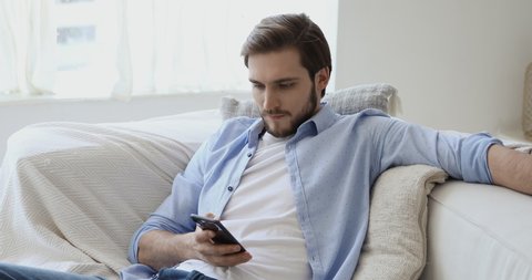 Handsome young european appearance man relaxing on comfortable couch, using mobile application in living room. Happy caucasian guy chatting in social network, dating online or web surfing internet.