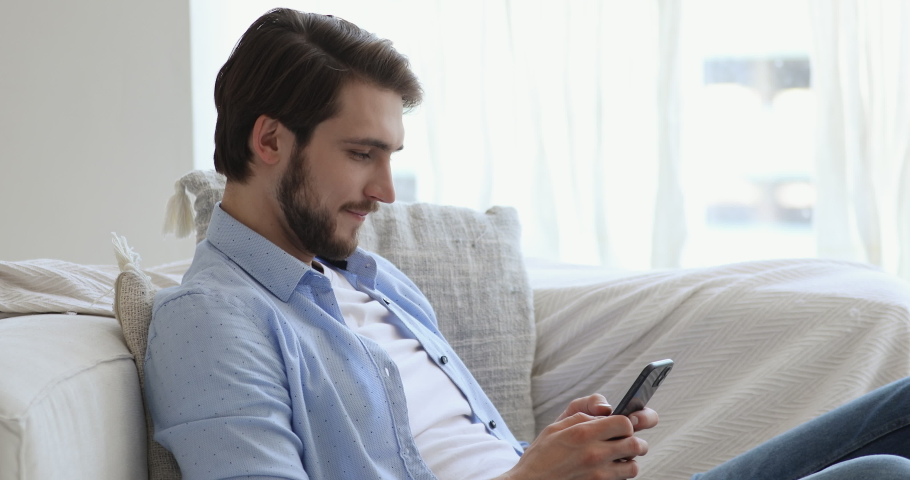 Side view happy young handsome guy sitting on sofa, using dating online mobile application indoors. Smiling man enjoying communicating with friends in social network or playing games on phone. | Shutterstock HD Video #1057121849