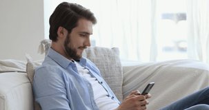 Side view happy young handsome guy sitting on sofa, using dating online mobile application indoors. Smiling man enjoying communicating with friends in social network or playing games on phone.