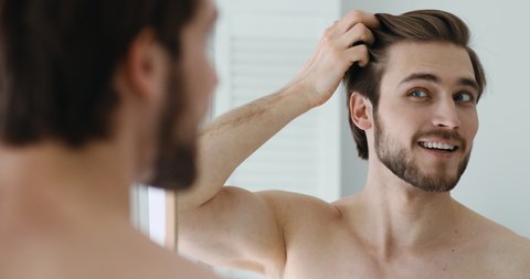 Smiling shirtless caucasian man looking at mirror, combing hair with fingers, getting ready for new day in bathroom. Head shot close up happy young stylish guy enjoying morning beauty routine indoors.
