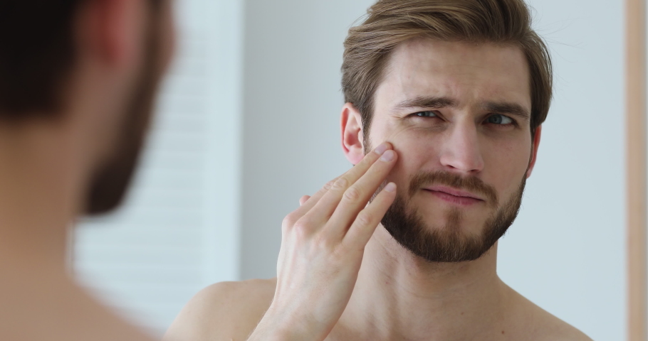 Close up head shot mirror reflection young man annoyed with dry unhealthy skin look, worrying about facial problems. Unhappy guy touching cheek, feeling stressed on oily skin or acne in bathroom. | Shutterstock HD Video #1057121894