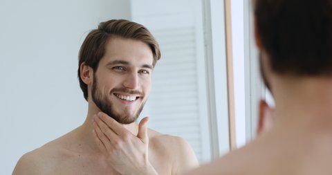 Mirror reflection young handsome smiling man touching beard, grooming in bathroom. Head shot close up happy confident shirtless european guy doing morning beauty routine, preparing for working day.