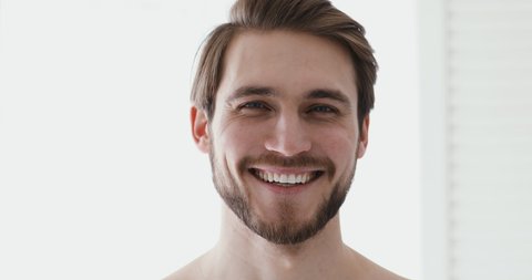 Close up head shot portrait young smiling handsome european appearance man standing in bathroom. Healthy bearded man with healthy white toothy smile looking at camera, feeling energetic in morning.