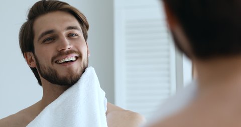 Mirror reflection young happy handsome bare man wiping face with towel, feeling energetic after shower in morning. Smiling well-groomed guy getting ready, doing morning skincare routine in bathroom.