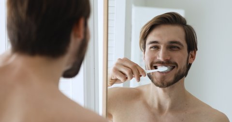 Close up head shot mirror reflection young happy bare man brushing teeth with toothbrush and toothpaste. Smiling handsome guy enjoying morning oral anti cavity hygienic routine alone in bathroom.